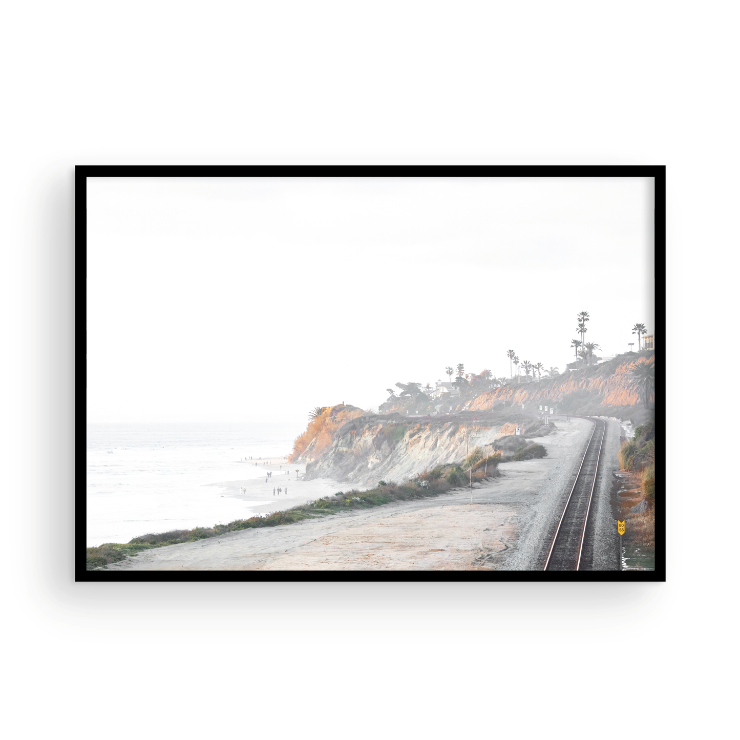 A slightly hazy, wide Landscape Coastal Photography shot shows a set of train tracks curving along a coastal cliffside, San Diego Coast. To the left, a sandy beach meets a light blue ocean waves with gentle waves, where a few small figures of people are visible. The cliffs are varied in color, with sections of tan, white, and hints of orange, topped with greenery and a few Palm Trees. The sky is a muted white, perfect for Minimalist Surf Wall Art. Photograph in minimalist mock up