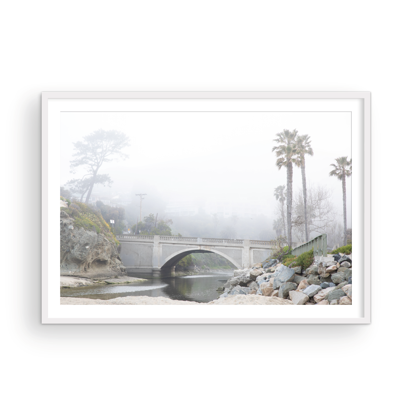 Landscape coastal photography of a concrete arch bridge disappearing into a dense fog, flanked by palm trees and rocky shores. This calm beach print evokes a mysterious seascape. Photograph in minimalist mock up.