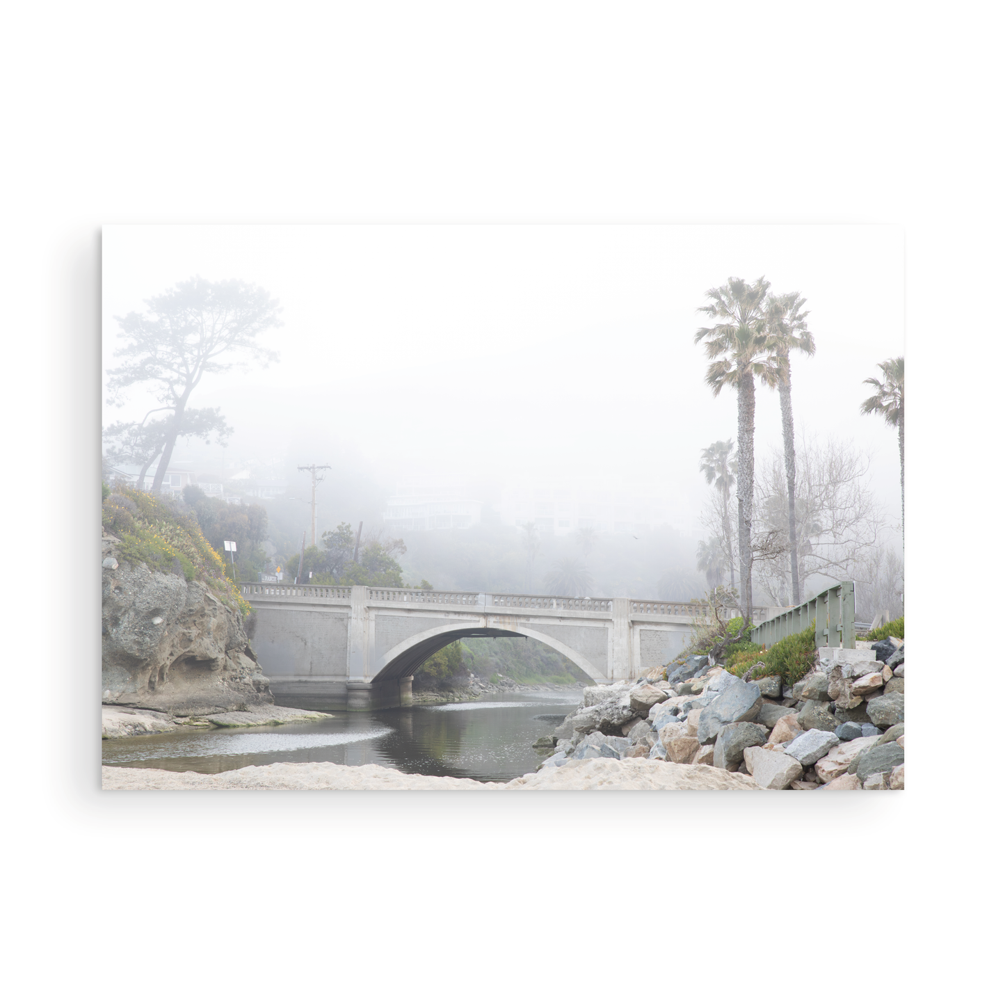 Landscape coastal photography of a concrete arch bridge disappearing into a dense fog, flanked by palm trees and rocky shores. This calm beach print evokes a mysterious seascape. Photograph in minimalist mock up.