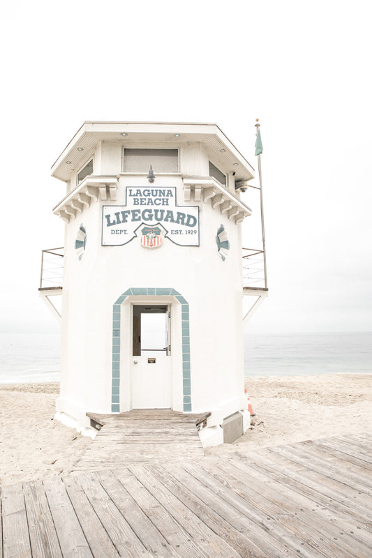 Vertical coastal photography of a white lifeguard tower at Laguna Beach, California, with a clear blue sky and ocean in the background. This neutral ocean print highlights coastal architecture. Photograph in minimalist mock up.