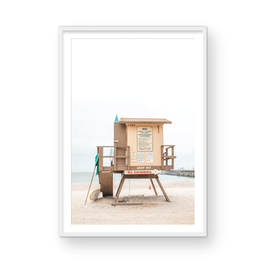 A vertical beach art close-up of a rustic lifeguard tower on San Clemente Beach, with a surfboard leaning against it and the pier visible in the distance. This image is perfect for a minimalist surf wall decor, surf nursery decor, or a fun surf print. Photograph in minimalist mock up.