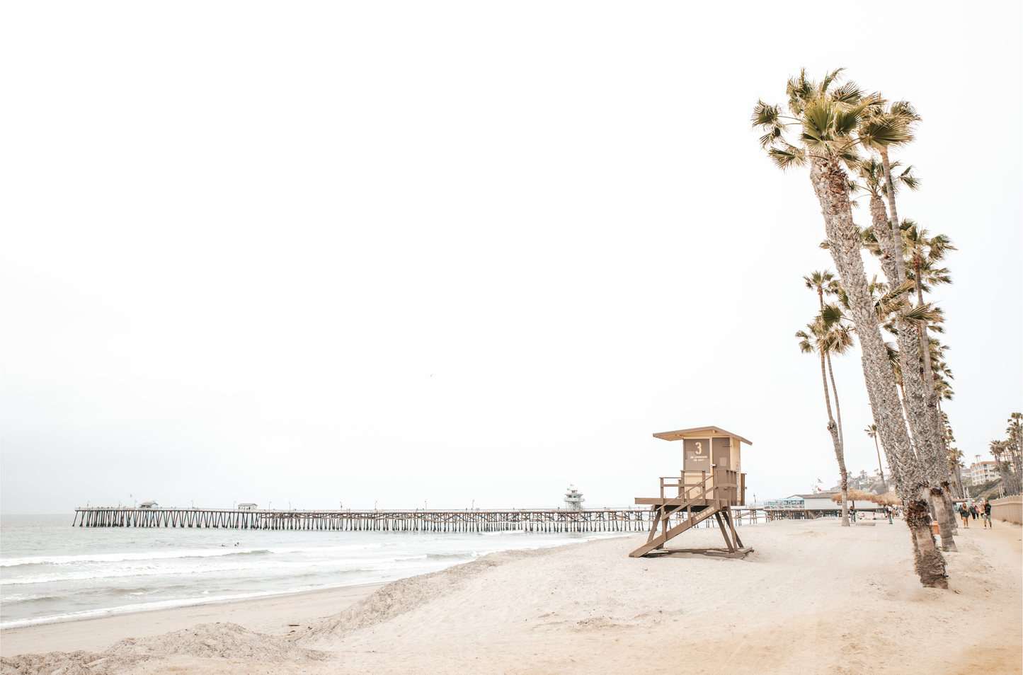 A wide landscape coastal photography view of San Clemente State Beach in Southern California, featuring a row of tall palm trees, a lifeguard tower, and people enjoying the calm beach. This image evokes Summer Vibes Wall Art and tropical beach print aesthetics.