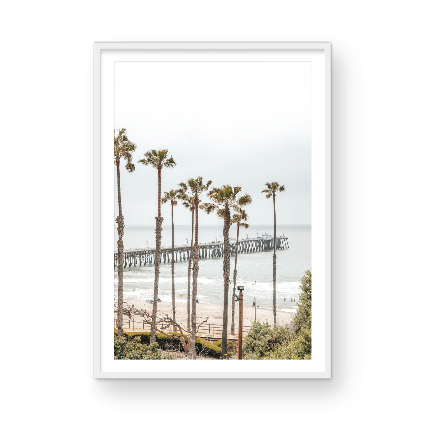 A vertical landscape coastal photography shot of the San Clemente Pier from behind a lush line of tall palm trees. This beachscape print highlights the California coast and is ideal for large beach art or a beach theme wall decor, with subtle ocean waves. Photograph in minimalist mock up.