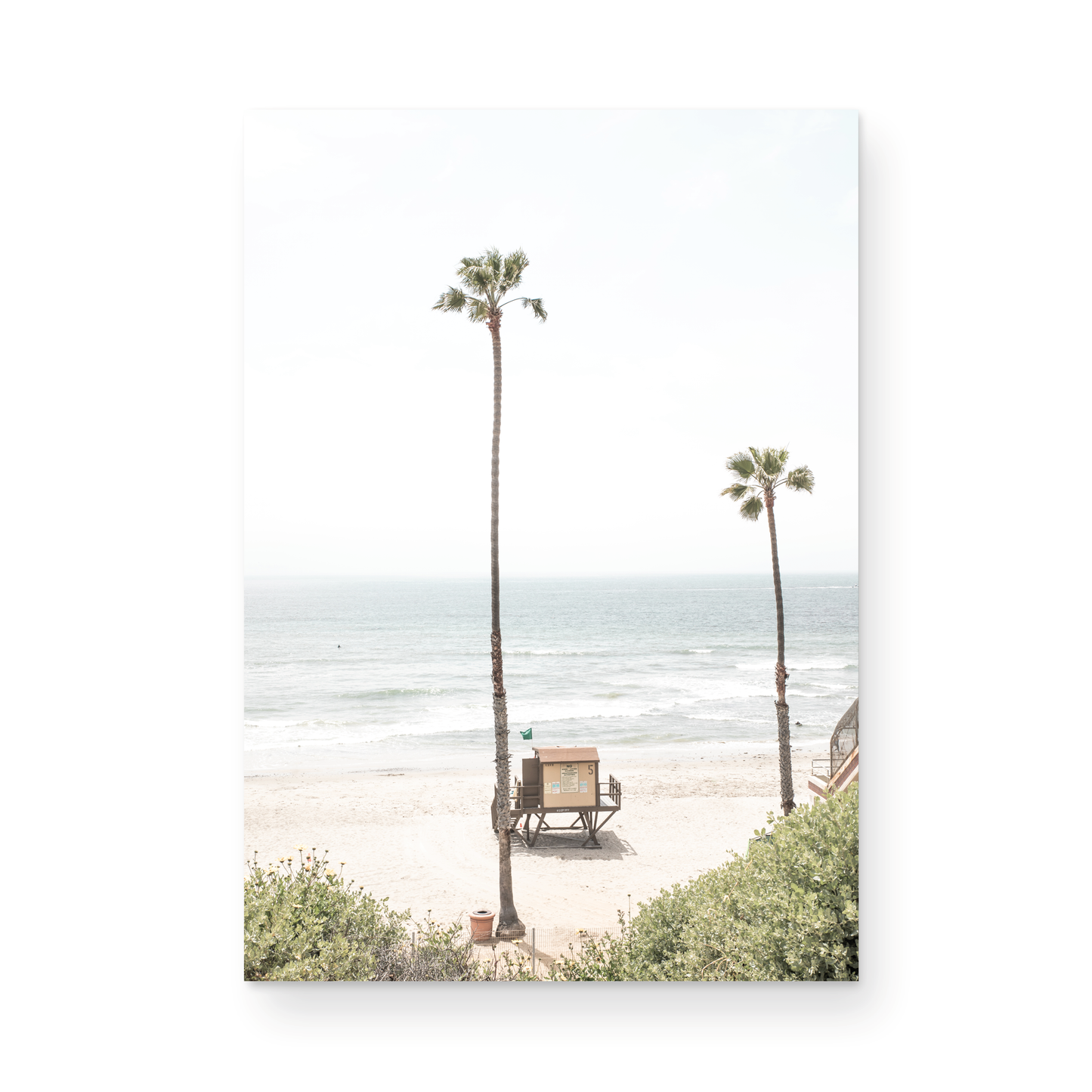 Vertical beach wall art featuring a lifeguard tower nestled between two tall palm trees on a sunny California beach, capturing a serene beach vibe. Photograph in minimalist mock up.