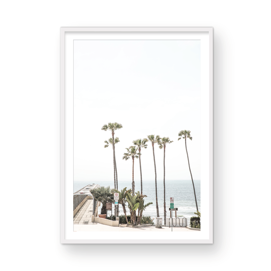 Vertical California coast print featuring a row of tall palm trees standing beside the entrance to a pier, with the ocean in the background. A coastal palm tree print. Photograph in minimalist mock up.