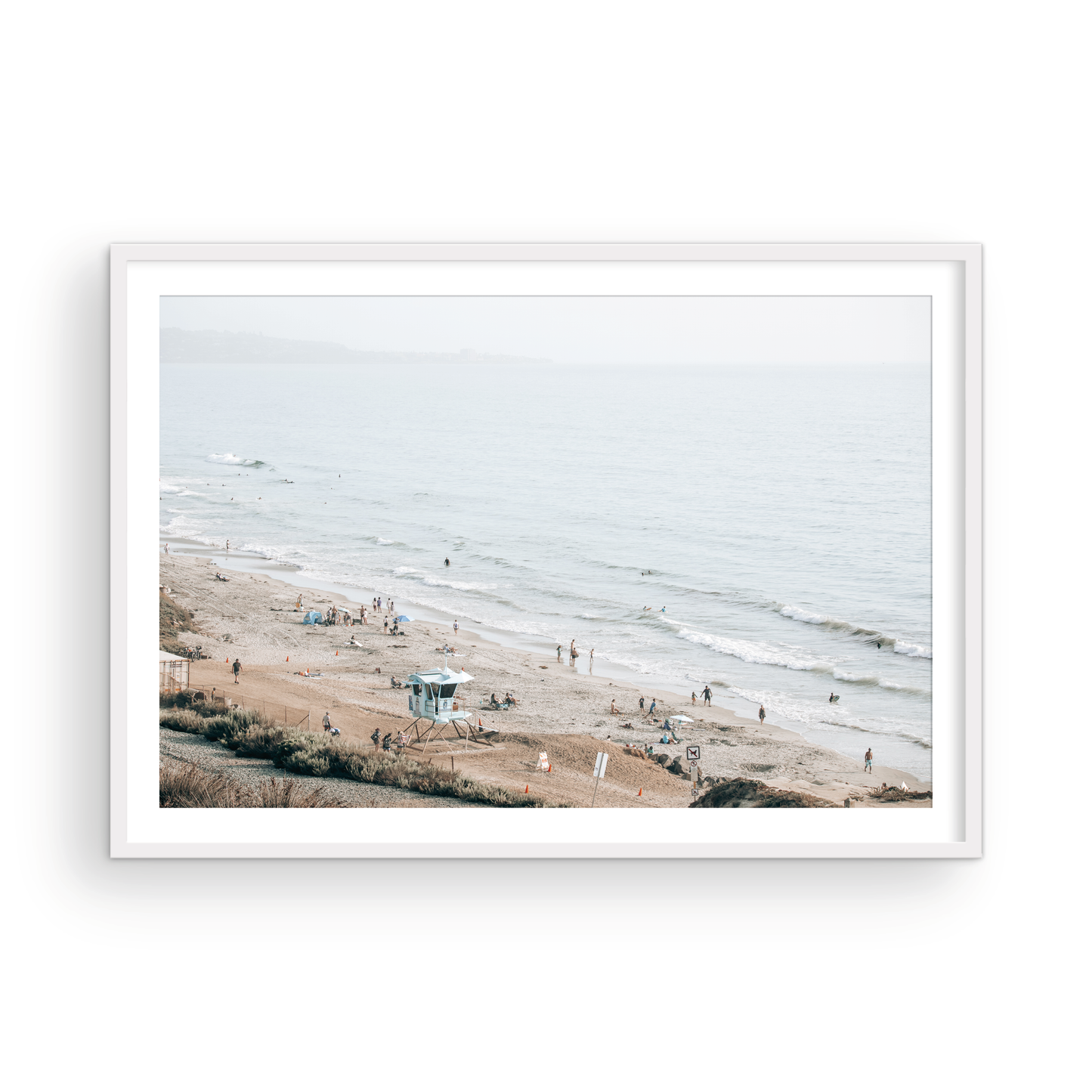 A slightly hazy, high-angle Landscape Beach Print captures a wide beachscape print where a light blue ocean waves meet a sandy shore dotted with numerous people enjoying the Summer Vibes. Gentle white waves roll towards the beach. A Blue Lifeguard Tower stands prominently in the lower center of the framed landscape beach print. The overall lighting suggests a bright, overcast travel photography day on the San Diego coast. Photograph in minimalist mock up