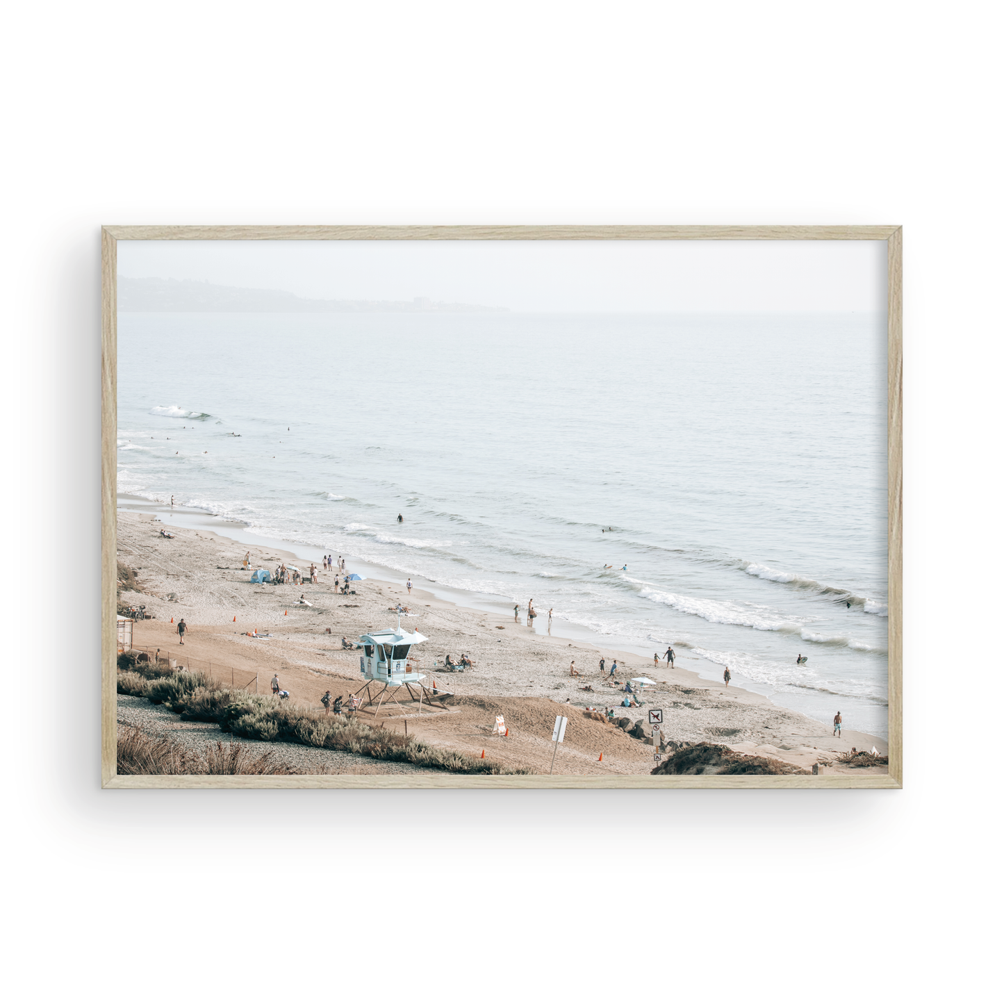 A slightly hazy, high-angle Landscape Beach Print captures a wide beachscape print where a light blue ocean waves meet a sandy shore dotted with numerous people enjoying the Summer Vibes. Gentle white waves roll towards the beach. A Blue Lifeguard Tower stands prominently in the lower center of the framed landscape beach print. The overall lighting suggests a bright, overcast travel photography day on the San Diego coast. Photograph in minimalist mock up