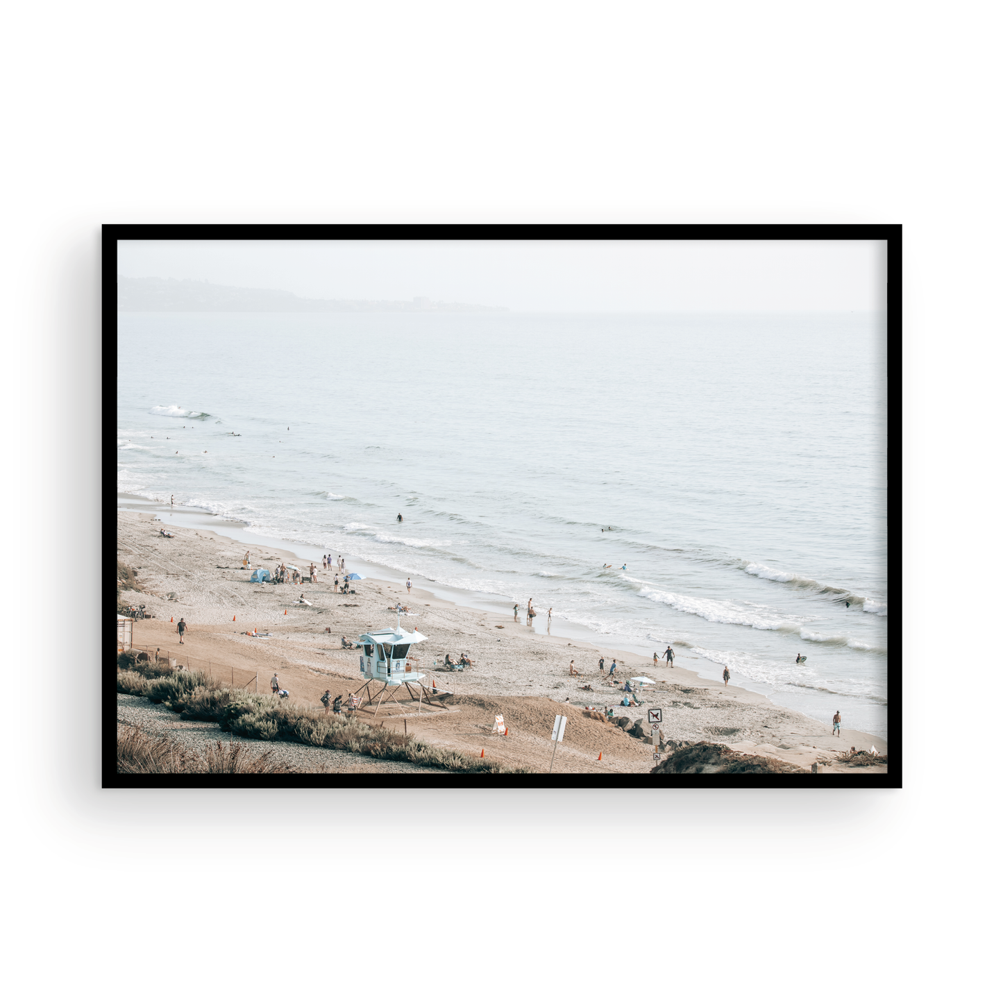 A slightly hazy, high-angle Landscape Beach Print captures a wide beachscape print where a light blue ocean waves meet a sandy shore dotted with numerous people enjoying the Summer Vibes. Gentle white waves roll towards the beach. A Blue Lifeguard Tower stands prominently in the lower center of the framed landscape beach print. The overall lighting suggests a bright, overcast travel photography day on the San Diego coast. Photograph in minimalist mock up