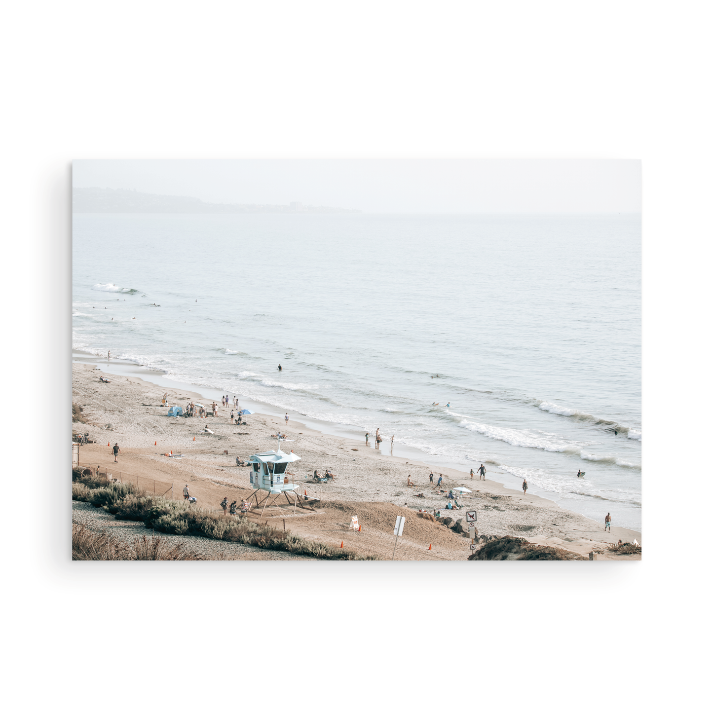 A slightly hazy, high-angle Landscape Beach Print captures a wide beachscape print where a light blue ocean waves meet a sandy shore dotted with numerous people enjoying the Summer Vibes. Gentle white waves roll towards the beach. A Blue Lifeguard Tower stands prominently in the lower center of the framed landscape beach print. The overall lighting suggests a bright, overcast travel photography day on the San Diego coast. Photograph in minimalist mock up