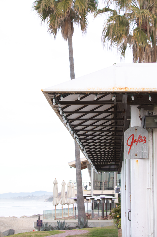 A framed beach scene art print with a wooden structure and palm trees, titled 'Jake's' on the sign, set against a background of a beach and ocean.