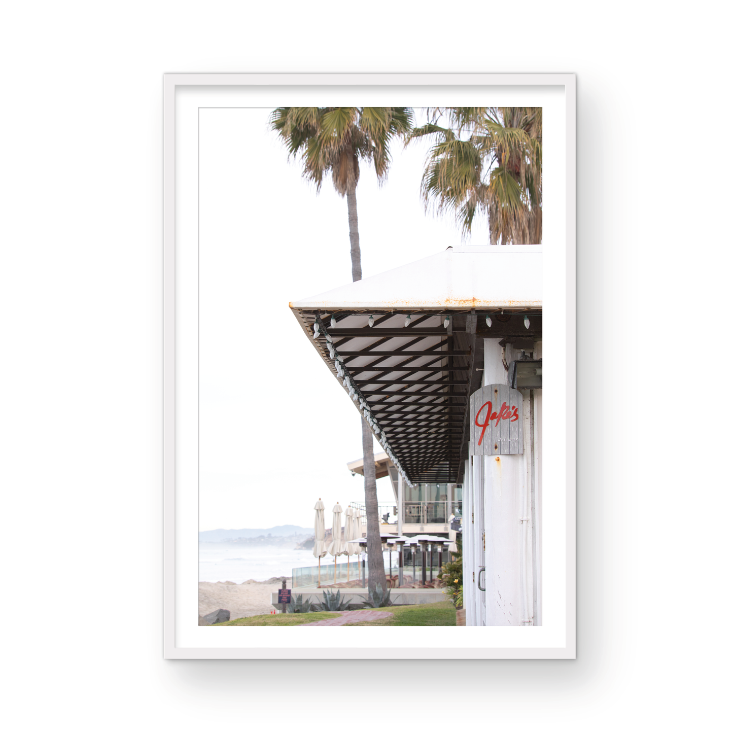 A framed beach scene art print with a wooden structure and palm trees, titled 'Jake's' on the sign, set against a background of a beach and ocean. Photograph in minimalist mock up.