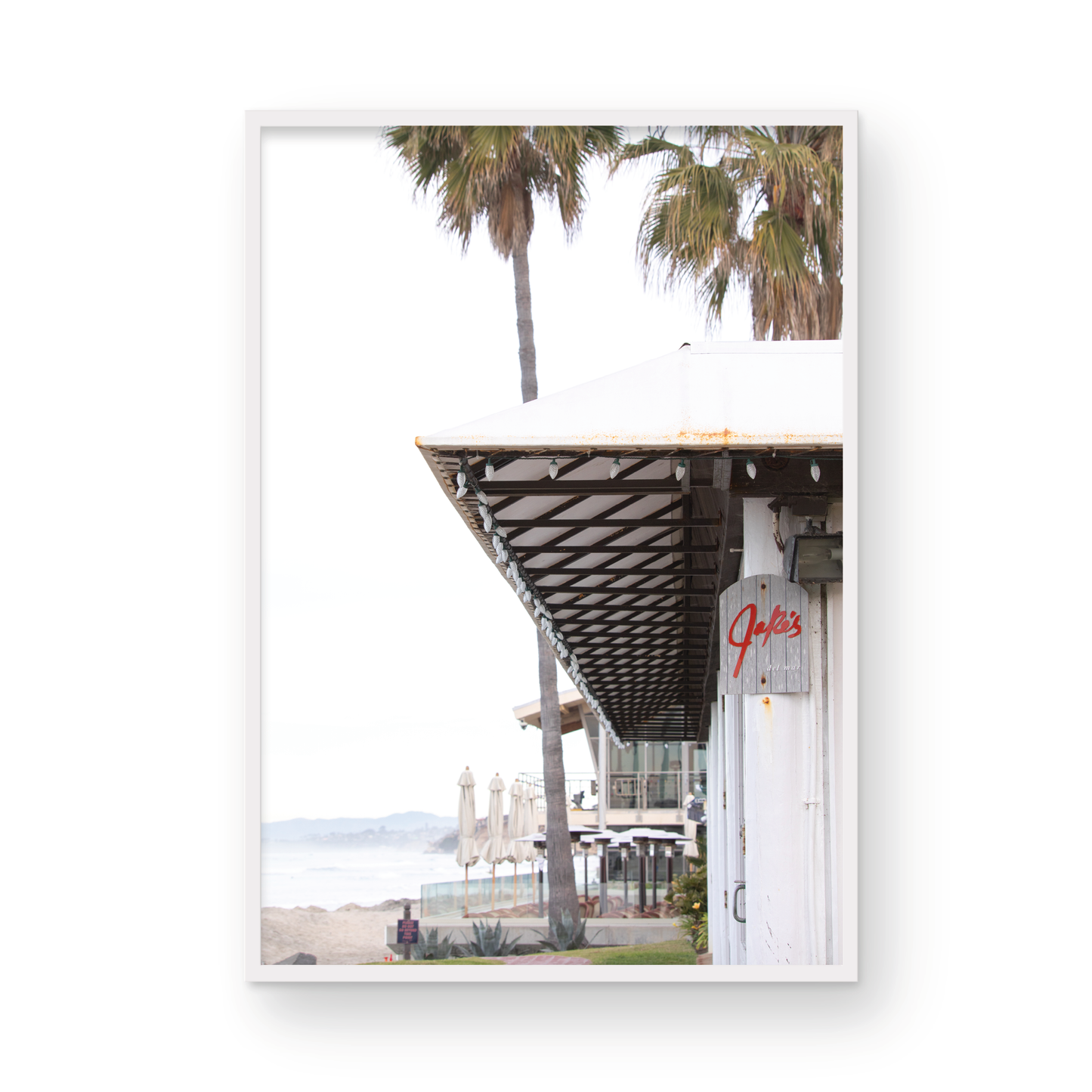 A framed beach scene art print with a wooden structure and palm trees, titled 'Jake's' on the sign, set against a background of a beach and ocean. Photograph in minimalist mock up.