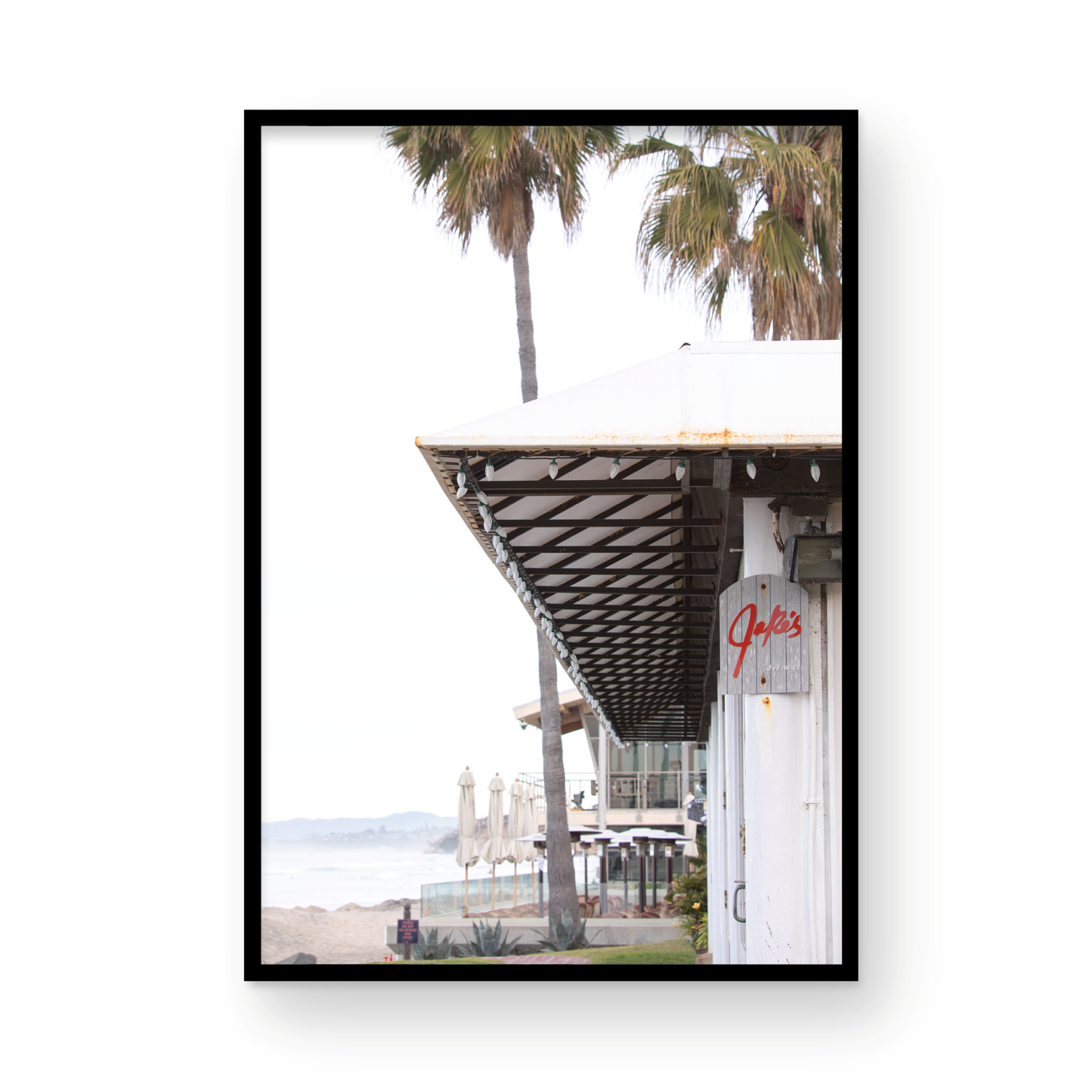 A framed beach scene art print with a wooden structure and palm trees, titled 'Jake's' on the sign, set against a background of a beach and ocean. Photograph in minimalist mock up.