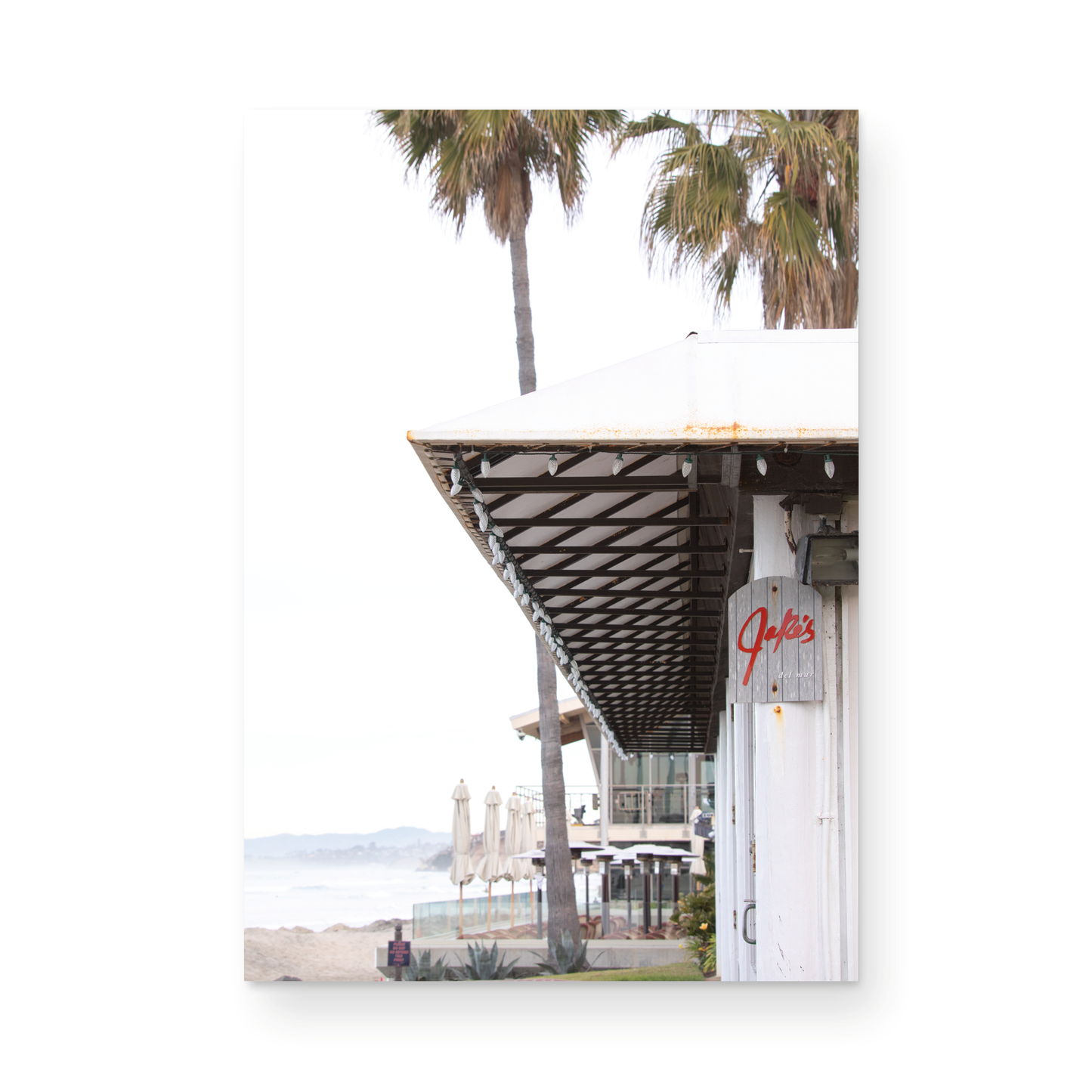 A framed beach scene art print with a wooden structure and palm trees, titled 'Jake's' on the sign, set against a background of a beach and ocean. Photograph in minimalist mock up.