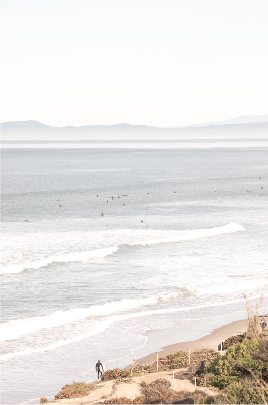 Minimalist ocean print of ocean waves with surfers in the distance and a sandy hillside in the foreground.