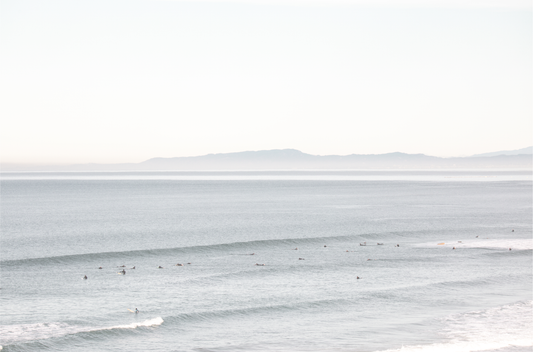 Coastal photography featuring a calm ocean scene with gentle waves and a hazy horizon line, predominantly in blue and white tones.