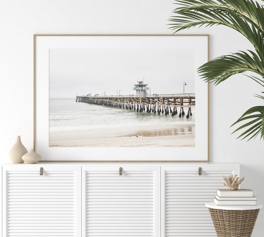 A long wooden pier, the San Clemente Pier, extends into the ocean in this landscape coastal photography. The beach in the foreground suggests a calm ocean print, ideal for a large coastal wall art or a beach cottage wall decor. Photograph in minimalist mock up.