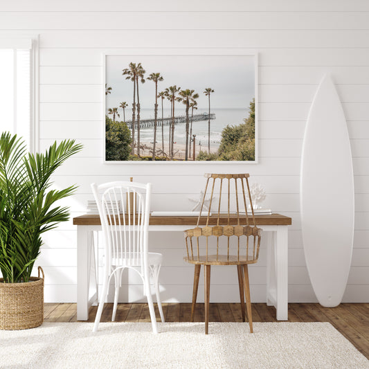 A horizontal landscape coastal photography shot of the San Clemente Pier from behind a lush line of tall palm trees. This beachscape print highlights the California coast and is ideal for large landscape beach art or a beach theme wall decor, with subtle ocean waves. Photograph in minimalist mock up.