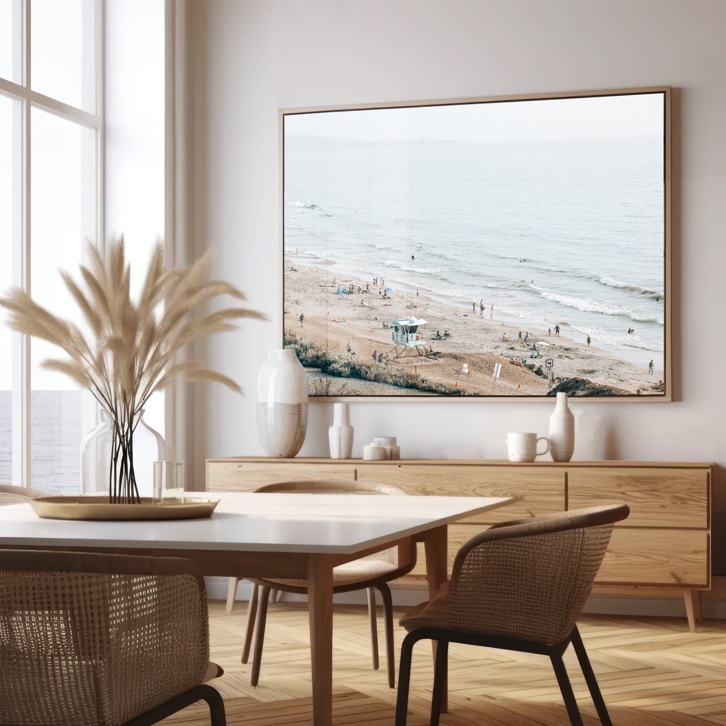A slightly hazy, high-angle Landscape Beach Print captures a wide beachscape print where a light blue ocean waves meet a sandy shore dotted with numerous people enjoying the Summer Vibes. Gentle white waves roll towards the beach. A Blue Lifeguard Tower stands prominently in the lower center of the framed landscape beach print. The overall lighting suggests a bright, overcast travel photography day on the San Diego coast. Photograph in minimalist dining room mock up