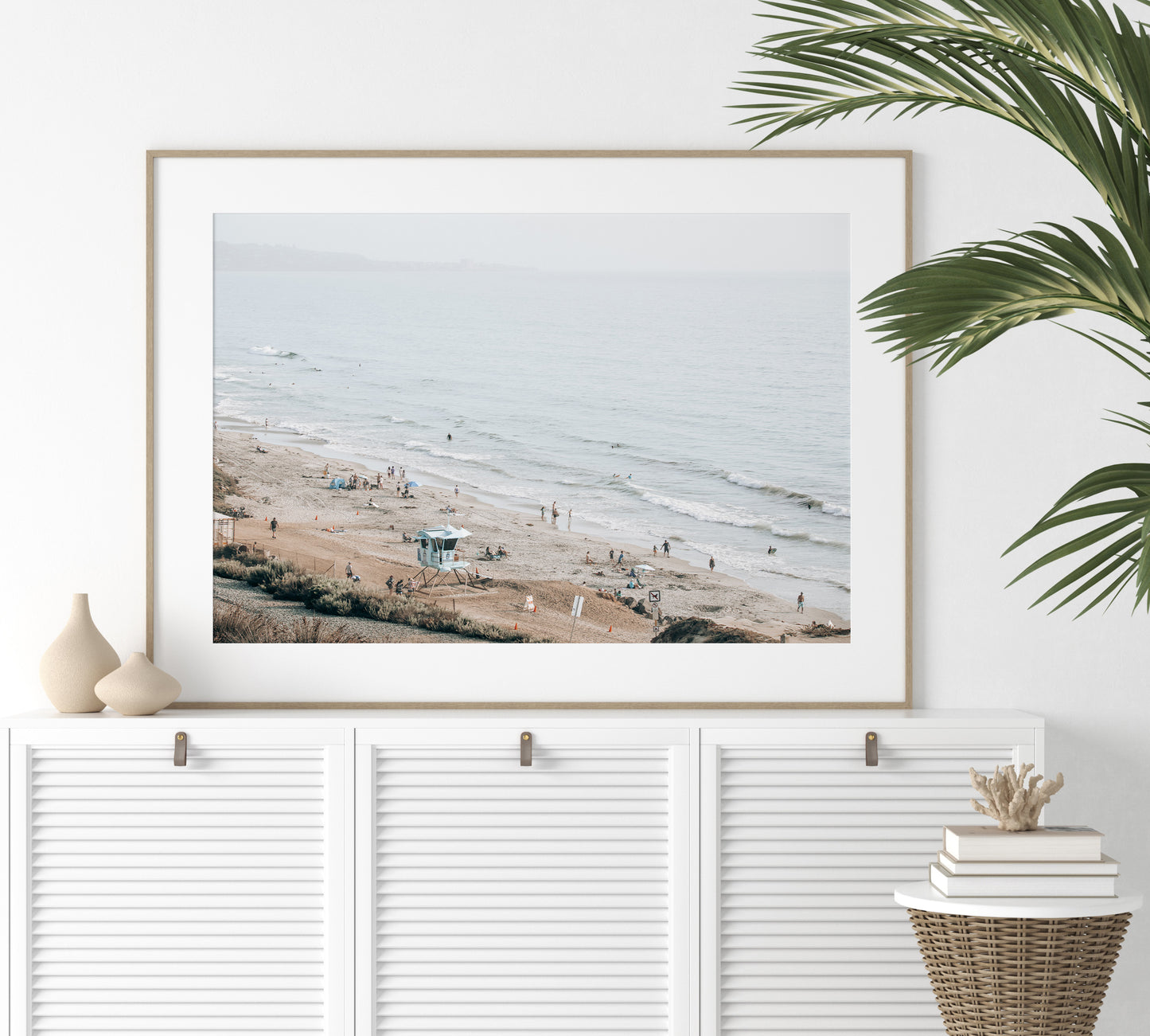 A slightly hazy, high-angle Landscape Beach Print captures a wide beachscape print where a light blue ocean waves meet a sandy shore dotted with numerous people enjoying the Summer Vibes. Gentle white waves roll towards the beach. A Blue Lifeguard Tower stands prominently in the lower center of the framed landscape beach print. The overall lighting suggests a bright, overcast travel photography day on the San Diego coast. Photograph in minimalist mock up
