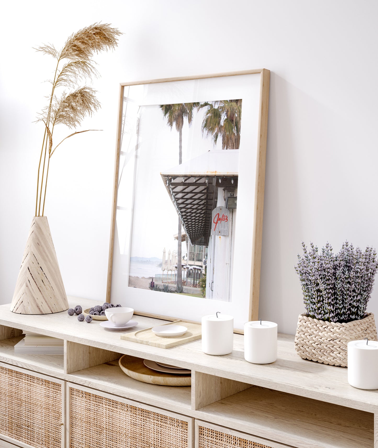 A framed beach scene art print with a wooden structure and palm trees, titled 'Jake's' on the sign, set against a background of a beach and ocean. Photograph in minimalist mock up.
