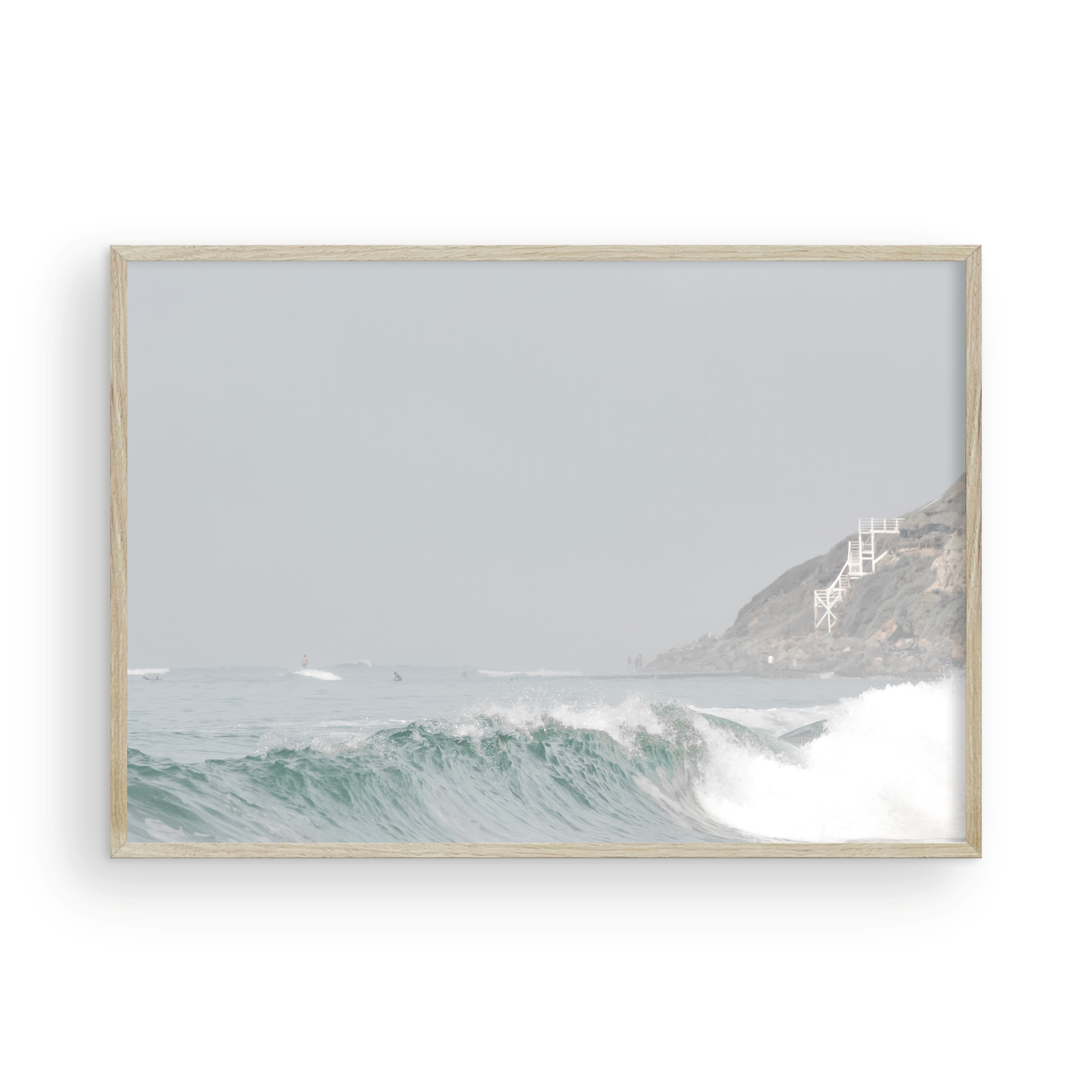 Neutral ocean print featuring a beautifully captured turquoise wave cresting with white seafoam, embodying minimalist surf art. This horizontal coastal photography showcases soft, pastel tones under a hazy sky. In the background, a rugged cliffside staircase on the right meets the sea, with faint distant surfers in the water, suggesting the expansive Southern California coastline. A tranquil piece of surf wall art, perfect for a coastal theme. Photograph in framed minimalist mock up.