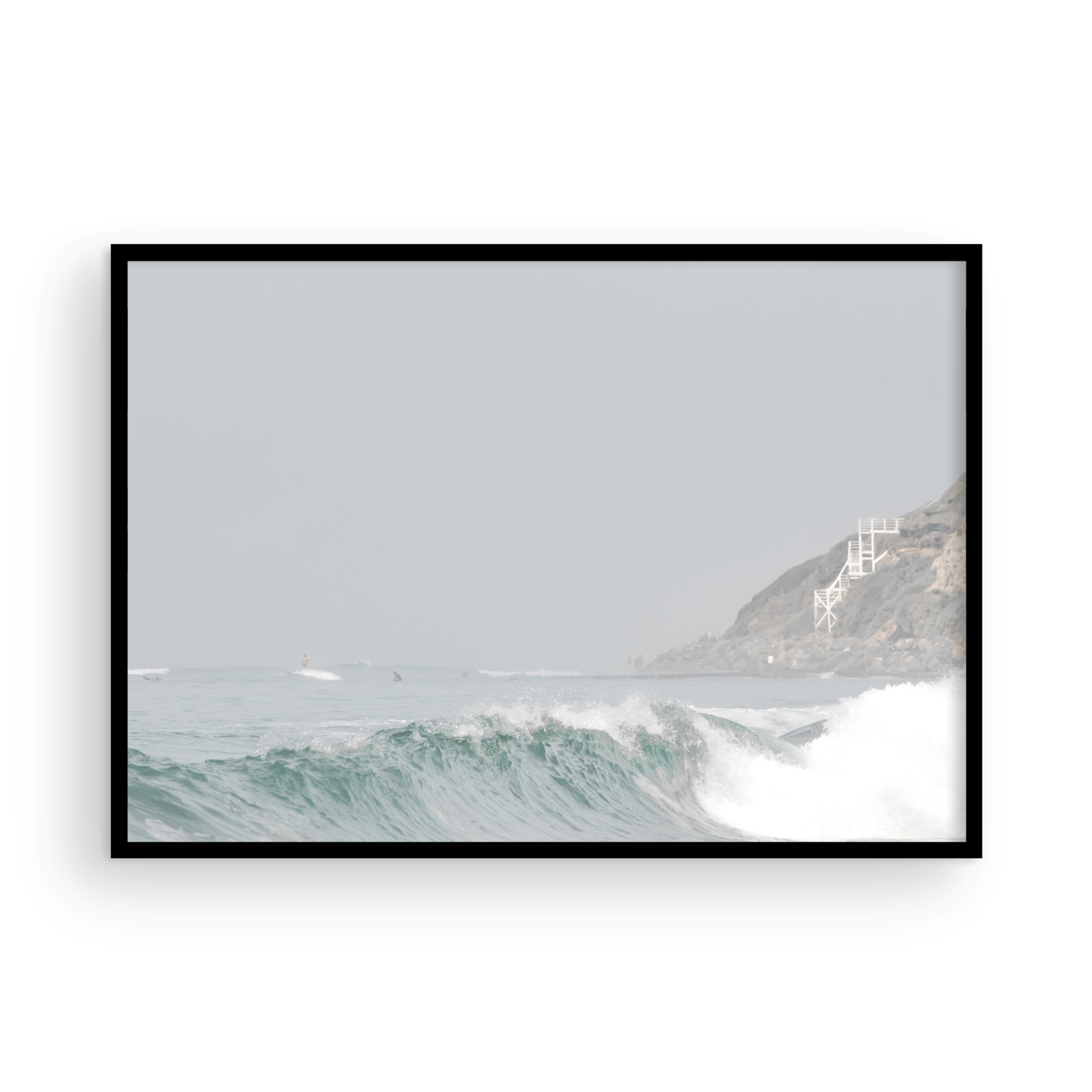 Neutral ocean print featuring a beautifully captured turquoise wave cresting with white seafoam, embodying minimalist surf art. This horizontal coastal photography showcases soft, pastel tones under a hazy sky. In the background, a rugged cliffside staircase on the right meets the sea, with faint distant surfers in the water, suggesting the expansive Southern California coastline. A tranquil piece of surf wall art, perfect for a coastal theme. Photograph in framed minimalist mock up.