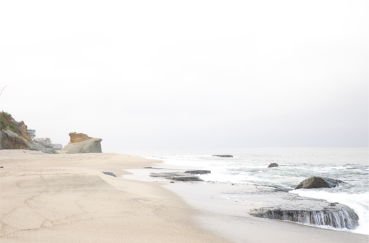 Horizontal coastal photography featuring a beach scene with calm ocean waves and a white sky, with rocks in the distance, located in Laguna California.