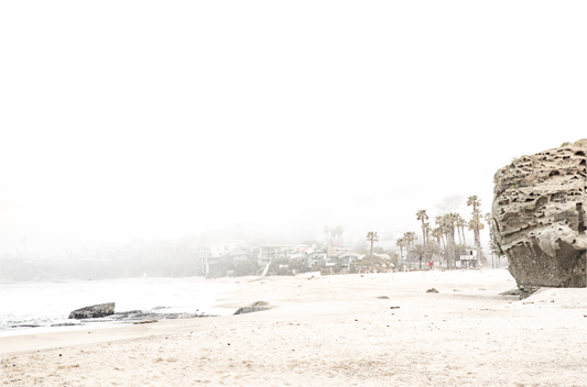 Horizontal coastal photography featuring a beach scene with a calm ocean and a rocky shore, beach homes in the foggy distance, located in Laguna California.