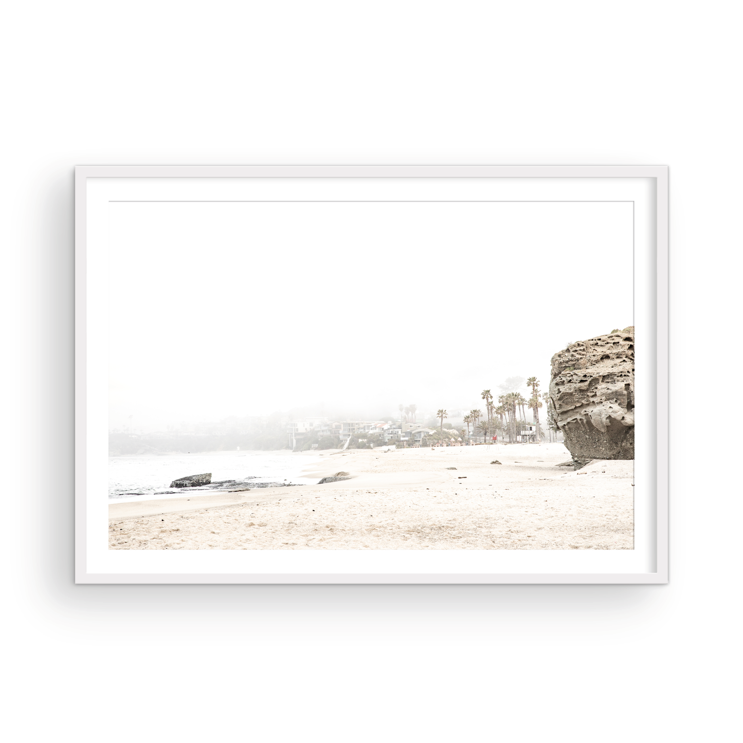 Horizontal coastal photography featuring a beach scene with a calm ocean and a rocky shore, beach homes in the foggy distance, located in Laguna California. Photograph in minimalist mock up.