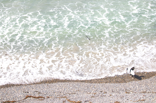 Horizontal surf print of a surfer with a surfboard walking along a rocky beach next to foamy ocean waves. This surfing art captures a unique coastline. Photograph in minimalist mock up.