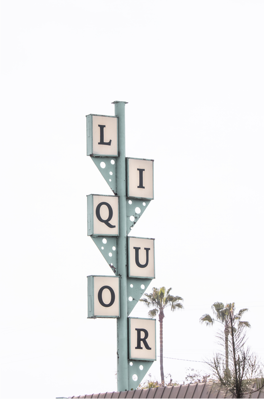 A vertical shot of the iconic Leucadia Liquor sign, with each letter of "LIQUOR" in a separate box against a teal-colored post and a bright white sky with palm trees in the background.