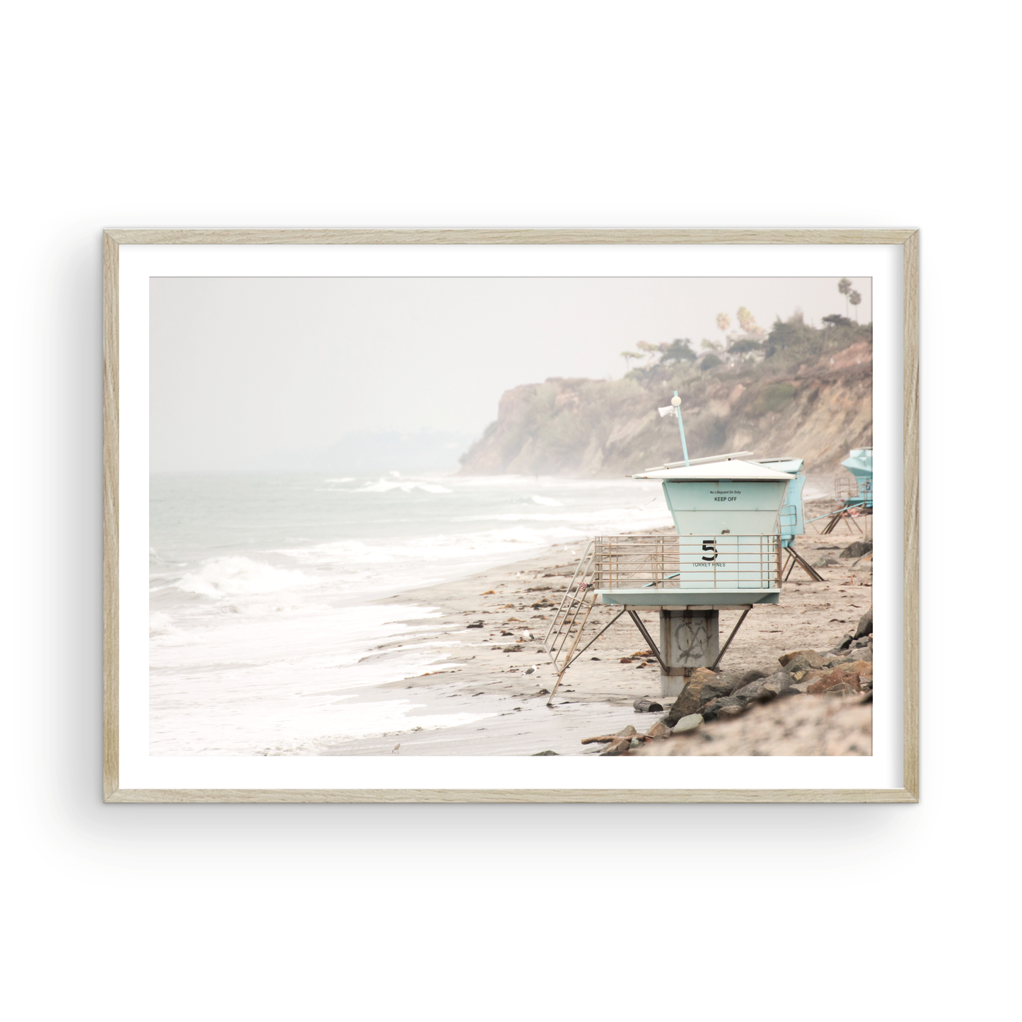 A serene piece of beach photography showcasing a light blue lifeguard tower, number 5, on the San Diego coast. This pastel beach art captures the iconic tower standing on wet sand with gentle ocean waves receding, under a hazy, overcast sky with distant cliffs. A perfect landscape lifeguard tower print for coastal theme wall art. Photograph in minimalist surf art mock up.