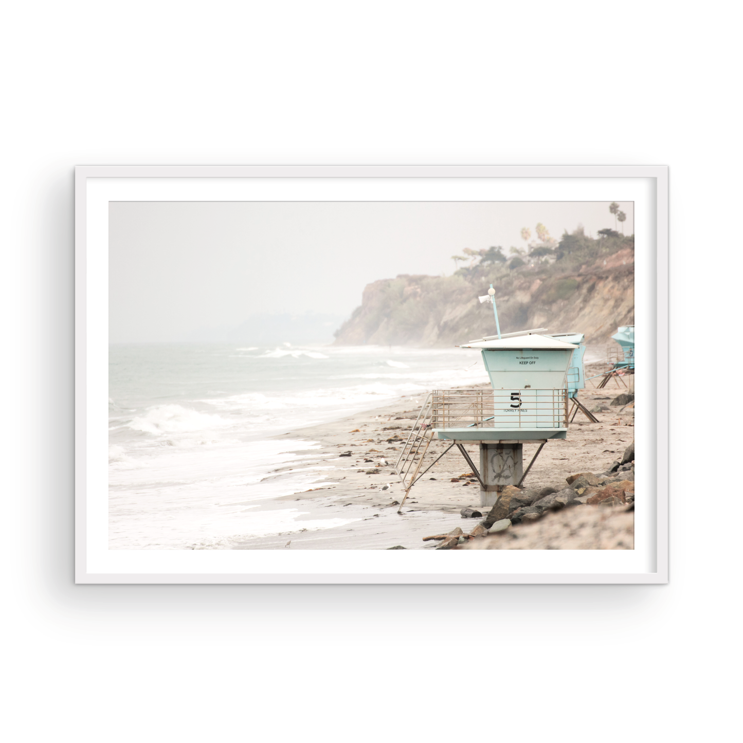 A serene piece of beach photography showcasing a light blue lifeguard tower, number 5, on the San Diego coast. This pastel beach art captures the iconic tower standing on wet sand with gentle ocean waves receding, under a hazy, overcast sky with distant cliffs. A perfect landscape lifeguard tower print for coastal theme wall art. Photograph in minimalist surf art mock up.