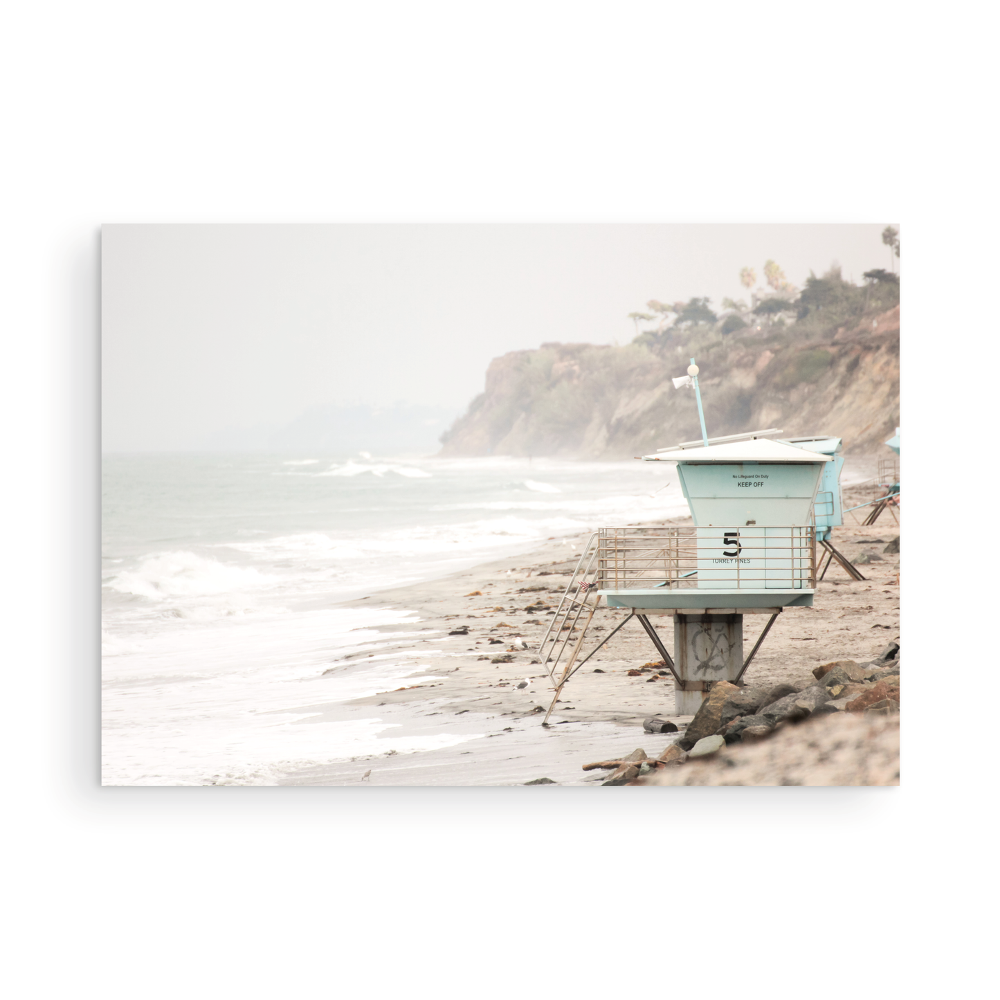 A serene piece of beach photography showcasing a light blue lifeguard tower, number 5, on the San Diego coast. This pastel beach art captures the iconic tower standing on wet sand with gentle ocean waves receding, under a hazy, overcast sky with distant cliffs. A perfect landscape lifeguard tower print for coastal theme wall art. Photograph in minimalist surf art mock up.