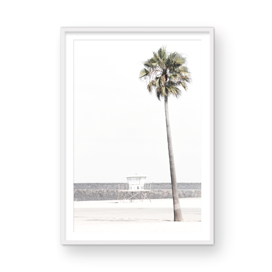 Dreamy coastal palm tree print featuring a tall palm tree in the foreground and a pastel blue lifeguard tower centered in the distance. This vertical tropical print is captured at Oceanside Beach on the Southern California coast, is bathed in bright, hazy sunlight, creating a pastel beach art effect. Photograph in minimalist mock up.