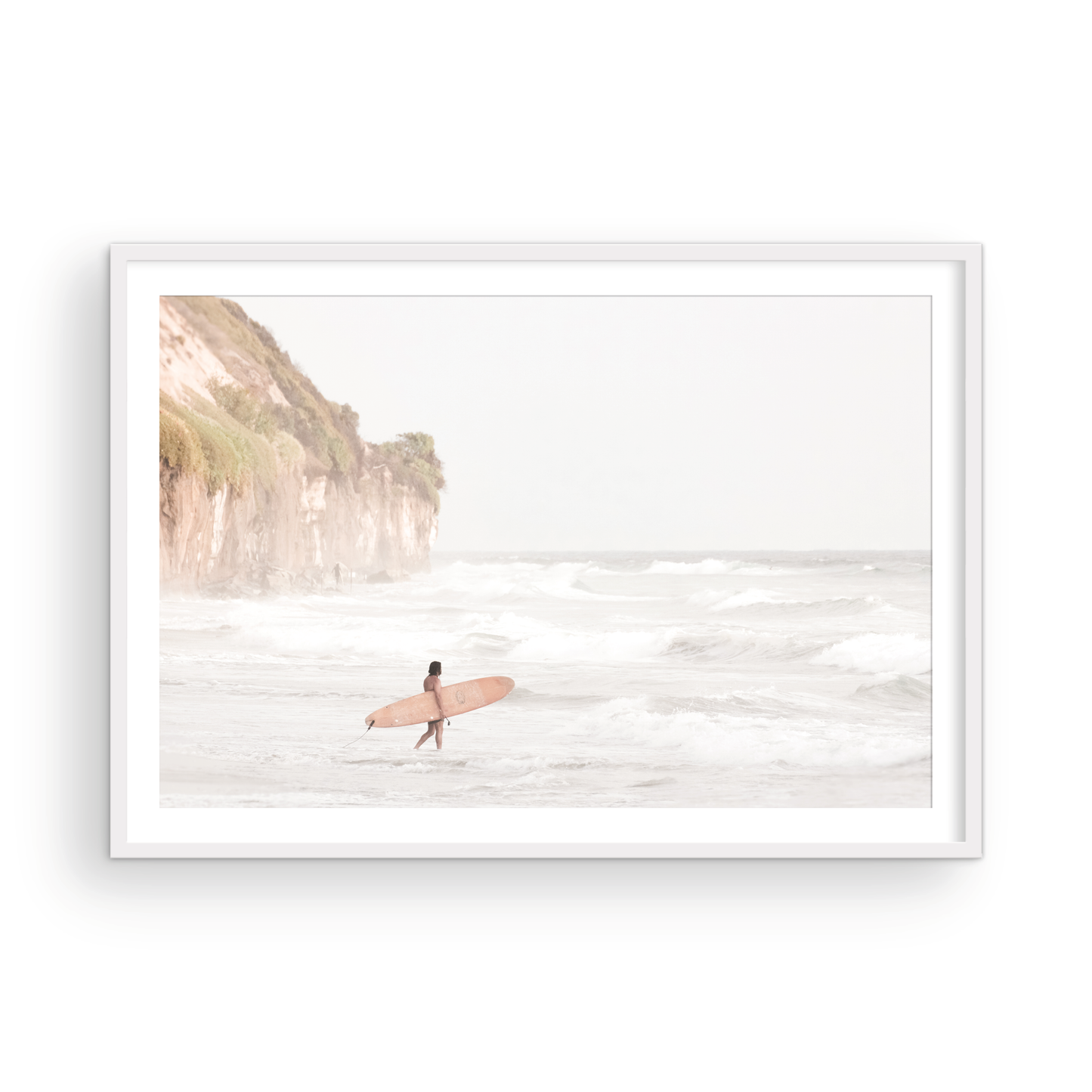 Horizontal surf art print of a lone surfer carrying a surfboard, walking on a rocky, foggy shoreline at Moonlight State Beach with a cliff in the background. Photograph in minimalist mock up.