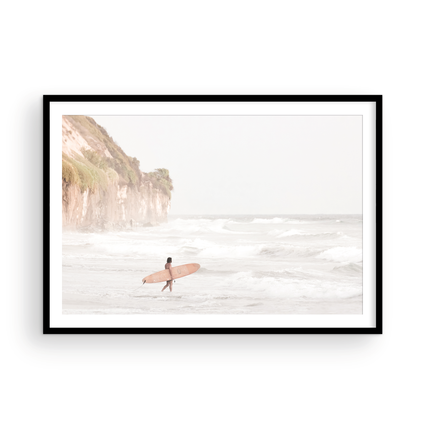 Horizontal surf art print of a lone surfer carrying a surfboard, walking on a rocky, foggy shoreline at Moonlight State Beach with a cliff in the background. Photograph in minimalist mock up.