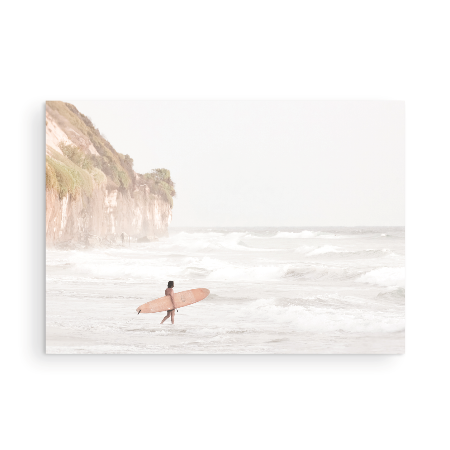 Horizontal surf art print of a lone surfer carrying a surfboard, walking on a rocky, foggy shoreline at Moonlight State Beach with a cliff in the background. Photograph in minimalist mock up.