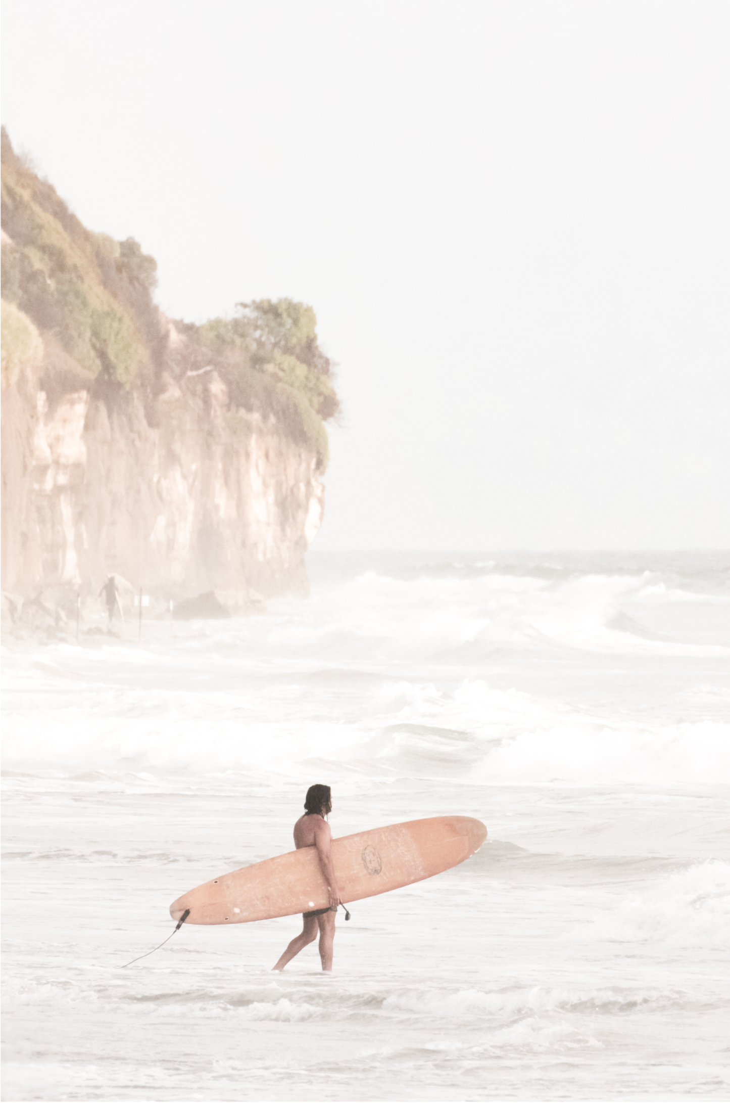 Vertical surf art print of a lone surfer carrying a surfboard, walking on a rocky, foggy shoreline at Moonlight State Beach with a lifeguard tower and cliff in the background.