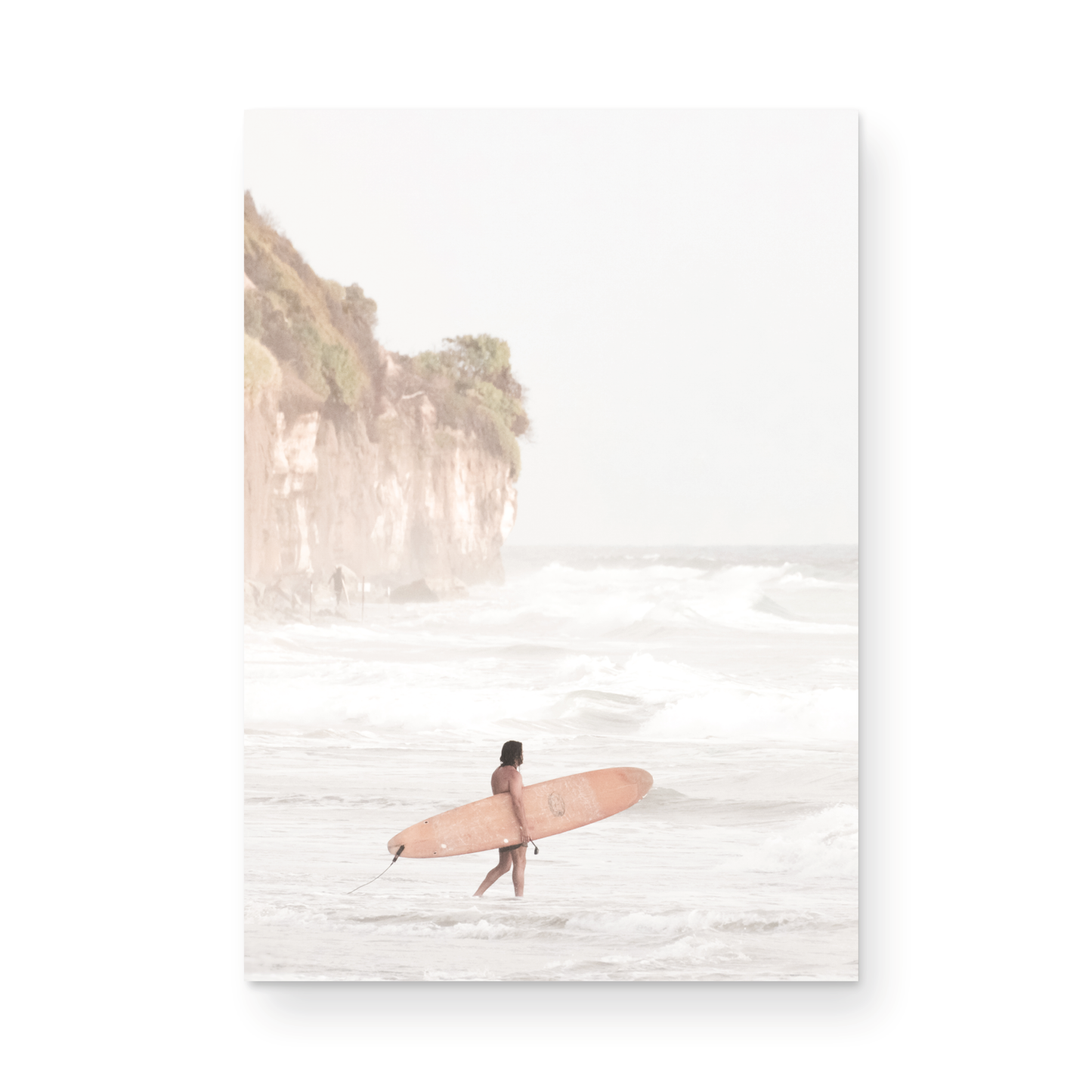 Vertical surf art print of a lone surfer carrying a surfboard, walking on a rocky, foggy shoreline at Moonlight State Beach with a lifeguard tower and cliff in the background. Photograph in minimalist mock up.