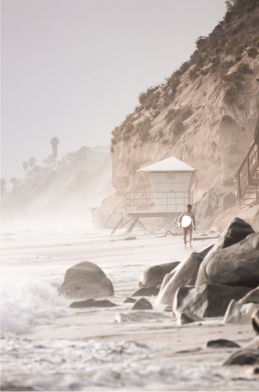 Vertical surf art print of a lone surfer carrying a surfboard, walking on a rocky, foggy shoreline at Moonlight State Beach with a lifeguard tower and cliff in the background.