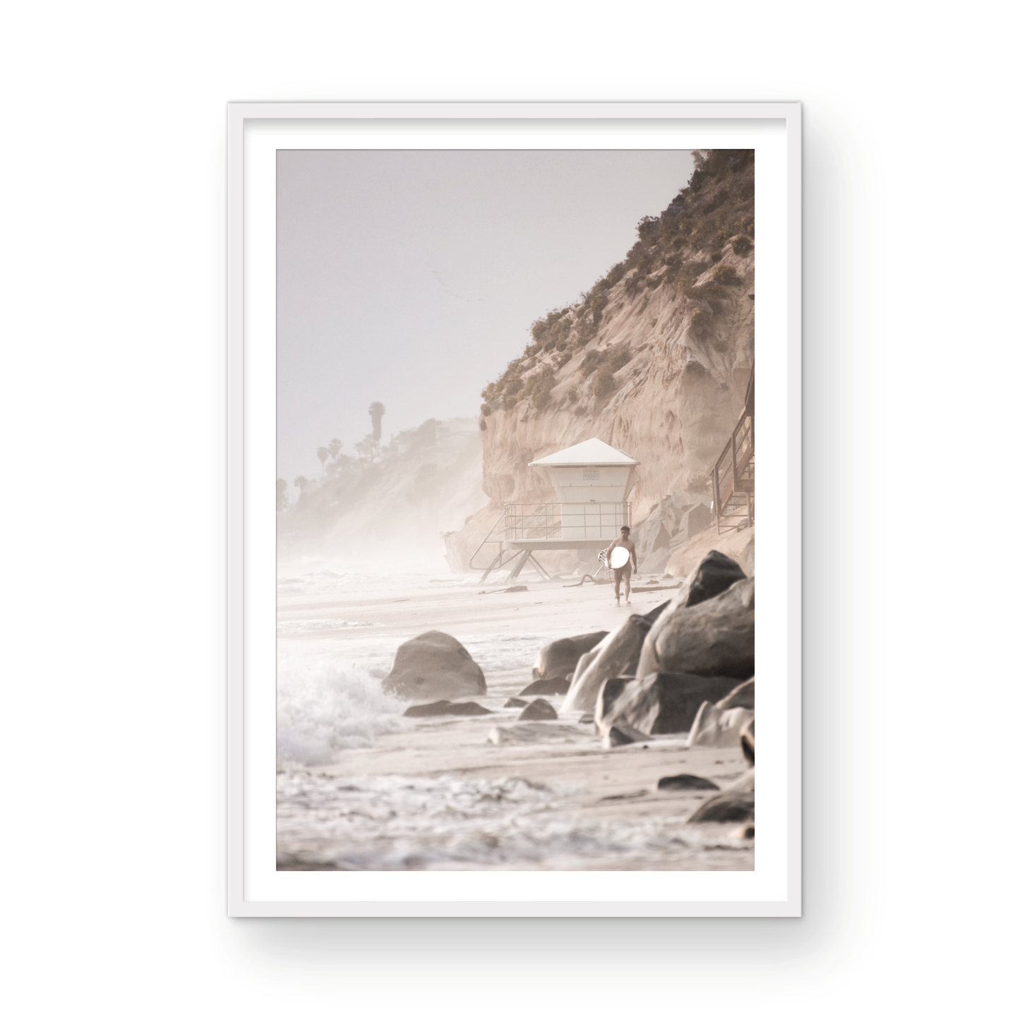Vertical surf art print of a lone surfer carrying a surfboard, walking on a rocky, foggy shoreline at Moonlight State Beach with a lifeguard tower and cliff in the background. Photograph in minimalist mock up.