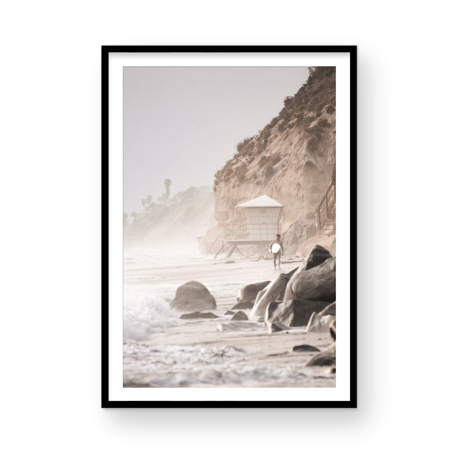 Vertical surf art print of a lone surfer carrying a surfboard, walking on a rocky, foggy shoreline at Moonlight State Beach with a lifeguard tower and cliff in the background. Photograph in minimalist mock up.