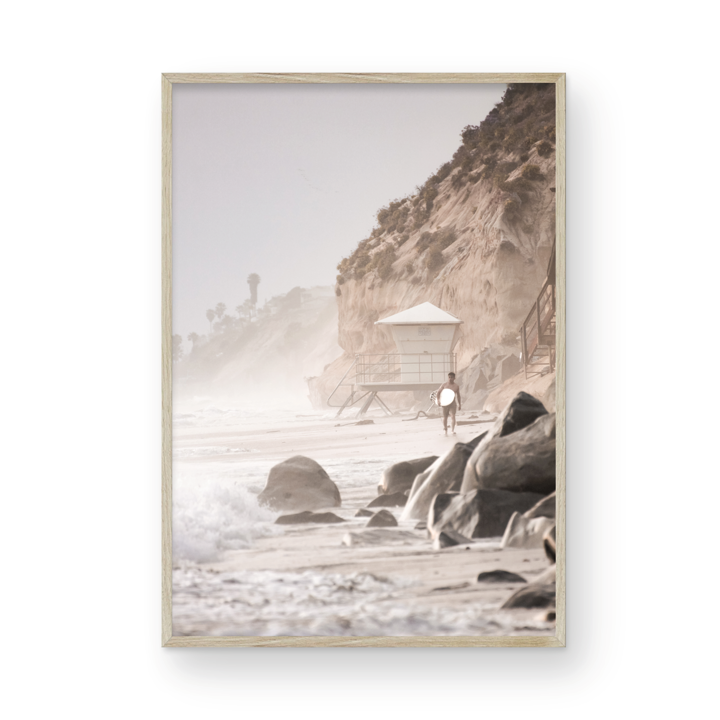 Vertical surf art print of a lone surfer carrying a surfboard, walking on a rocky, foggy shoreline at Moonlight State Beach with a lifeguard tower and cliff in the background. Photograph in minimalist mock up.