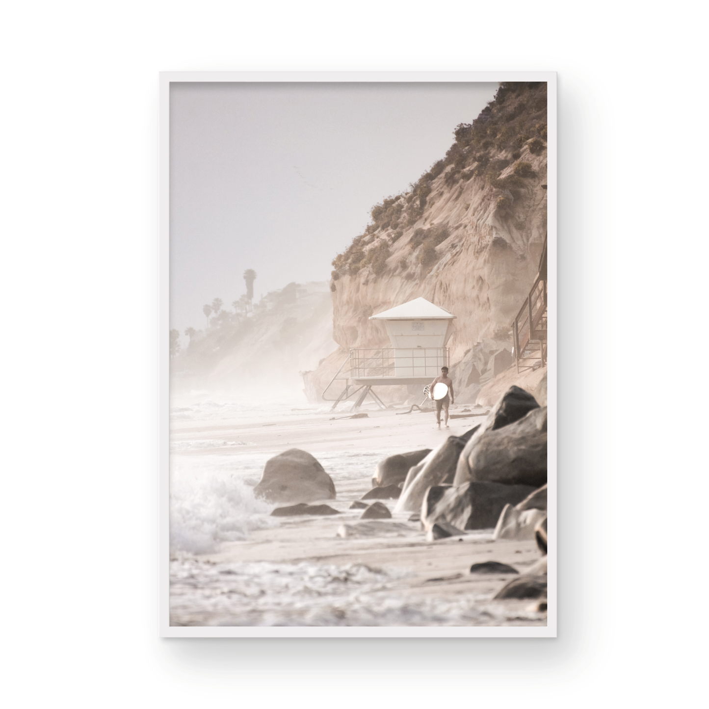 Vertical surf art print of a lone surfer carrying a surfboard, walking on a rocky, foggy shoreline at Moonlight State Beach with a lifeguard tower and cliff in the background. Photograph in minimalist mock up.