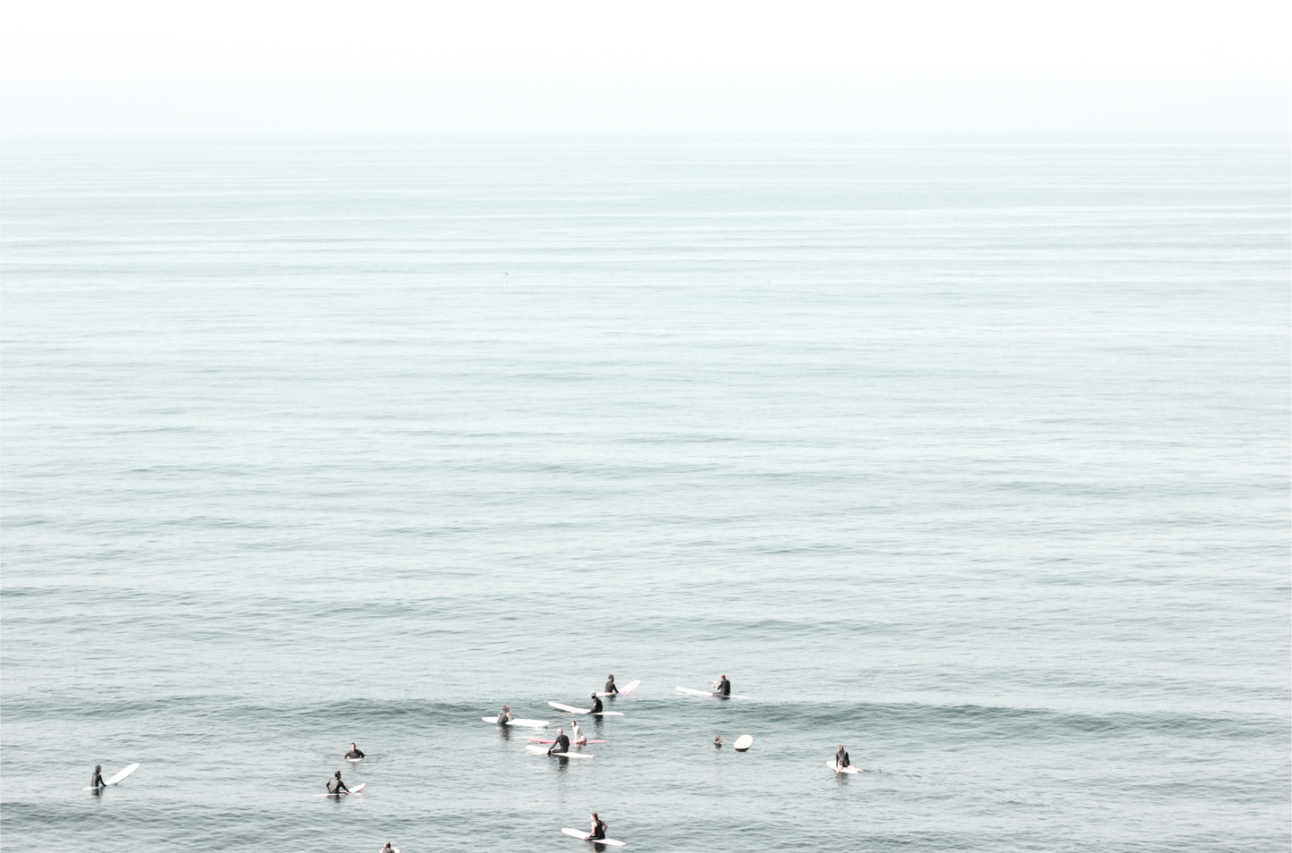 Horizontal surf art showing multiple surfers waiting in the calm ocean water for waves, with a hazy Southern California coastline in the distance. This minimalist surf print captures authentic surf vibes.