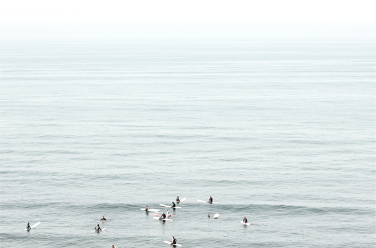 Horizontal surf art showing multiple surfers waiting in the calm ocean water for waves, with a hazy Southern California coastline in the distance. This minimalist surf print captures authentic surf vibes.