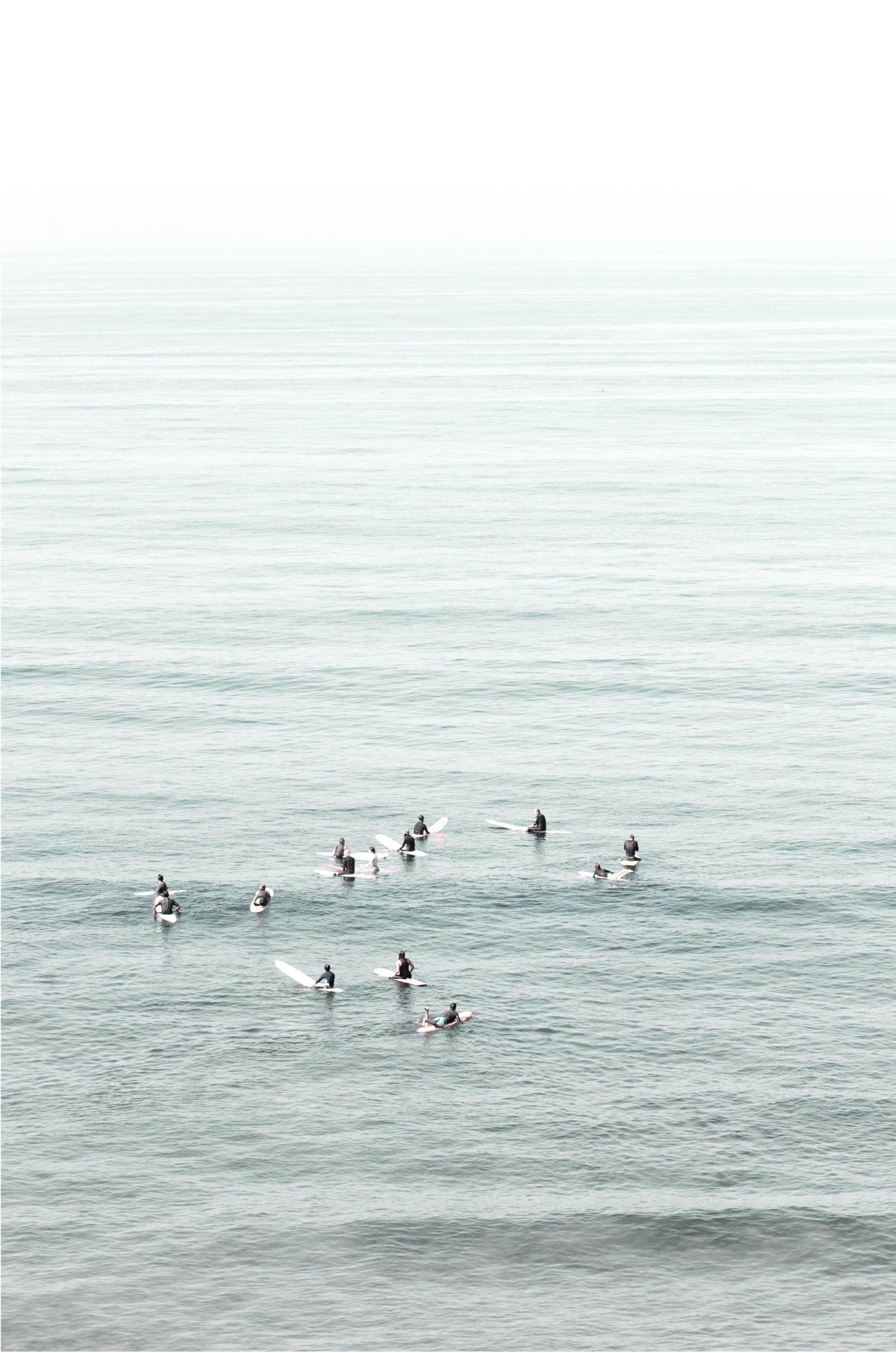 Vertical surf art showing multiple surfers waiting in the calm ocean water for waves, with a hazy Southern California coastline in the distance. This minimalist surf print captures authentic surf vibes.