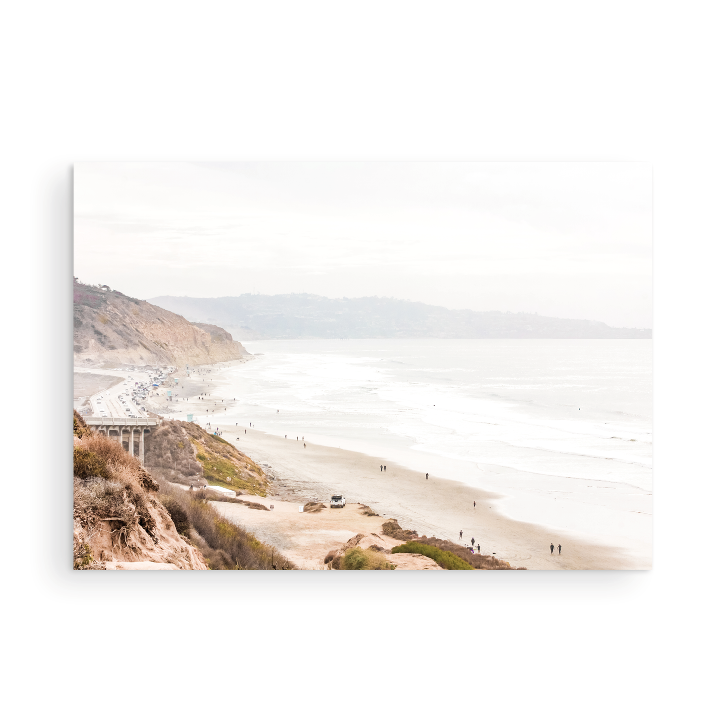 A slightly hazy, high-angle Landscape Beach Print captures a wide beachscape print where a light blue ocean waves meet a sandy shore dotted with numerous people enjoying the Summer Vibes. Gentle white waves roll towards the beach. Blue Lifeguard Towers line the coast in the far distance, of this framed landscape beach print. The overall lighting suggests a bright, overcast travel photography day on the San Diego coast. Photograph in minimalist mock up
